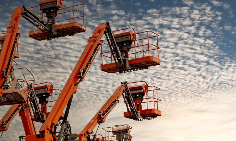 Plataformas elevatórias laranjas erguidas contra um céu azul com nuvens, prontas para uso em trabalhos em altura.